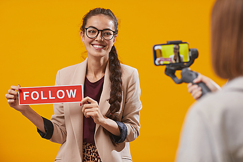 Waist up portrait of contemporary young woman holding Follow word while filming video for beauty and lifestyle channel standing against yellow background, copy space