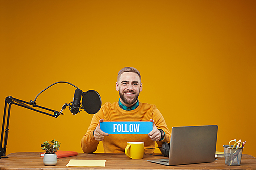 horizontal studio portrait of modern male working as video Vlog sitting at table holding plate with follow sign  smiling