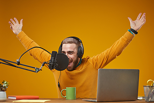 Horizontal medium studio portrait of joyful young Caucasian man wearing headphones recording content for his subscribers