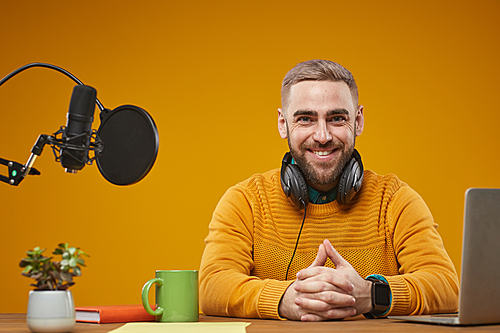 horizontal studio shot of handsome caucasian video Vlog wearing stylish mustard sweater sitting at desk  smiling
