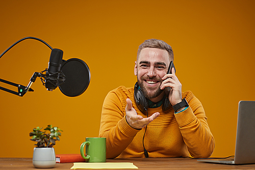 Horizontal studio portrait shot of young male radio presenter sitting at desk having phone call during break, mustard backround