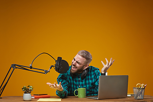 horizontal studio portrait of young male video Vlog giving emotional live stream show sitting at desk against mustard wall background