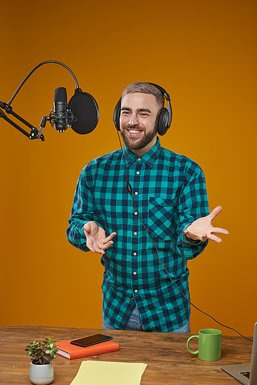 Vertical medium long shot portrait of young adult radio broadcaster wearing headphones recording content in his studio standing in front of microphone