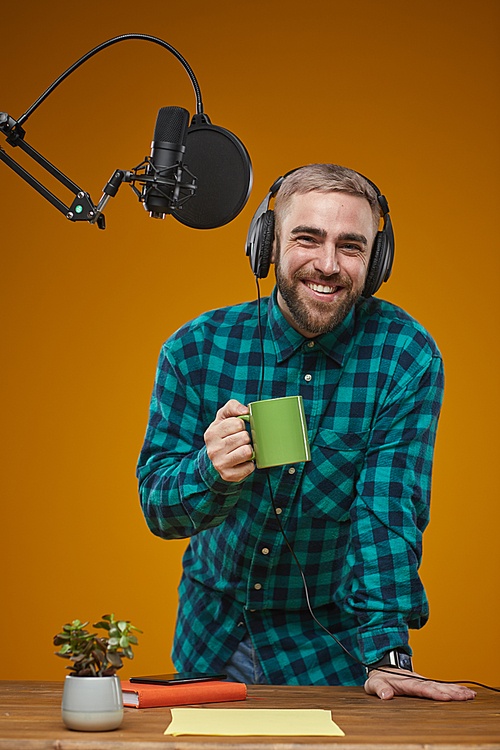 Vertical studio portrait of Caucasian radio presenter wearing checked shirt and headphones holding cup of coffee