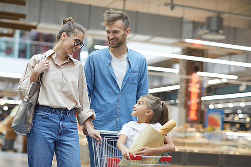 Portrait of happy young family grocery shopping together, parents smiling at little girl sitting in shopping cart, copy space