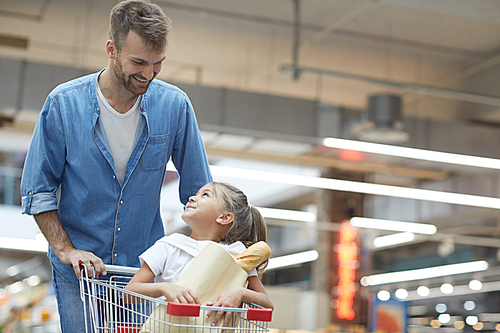 Portrait of happy young father grocery shopping in supermarket and smiling at little girl sitting in shopping cart, copy space