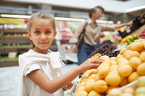 Waist up portrait of cute little girl choosing fresh fruits at farmers market and smiling at camera, copy space