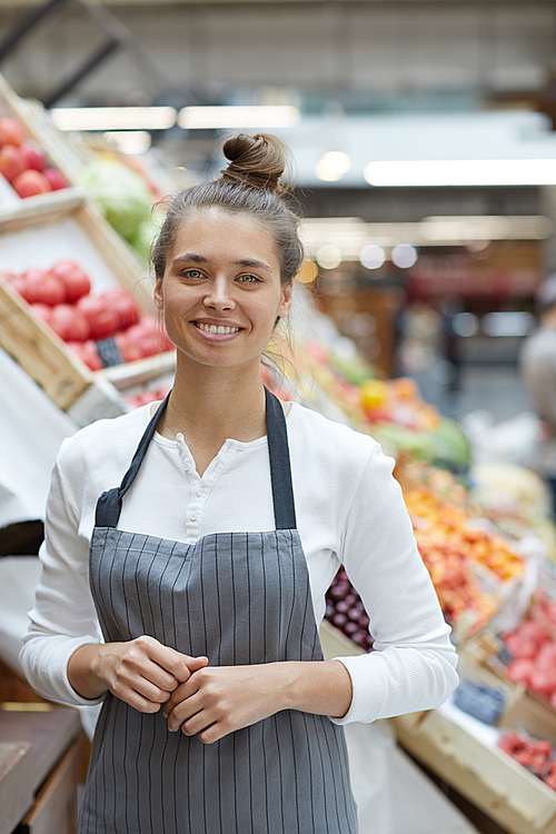 Waist up portrait of pretty young saleswoman smiling at camera while posing by fresh fruits and vegetables at supermarket, copy space