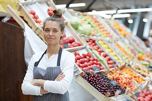 Waist up portrait of pretty young saleswoman  while posing by fresh fruits and vegetables at supermarket, copy space