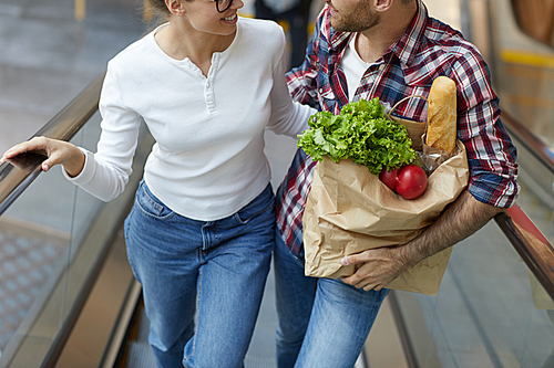 High angle portrait of contemporary young couple going up escalator in shopping mall and carrying grocery bag, copy space