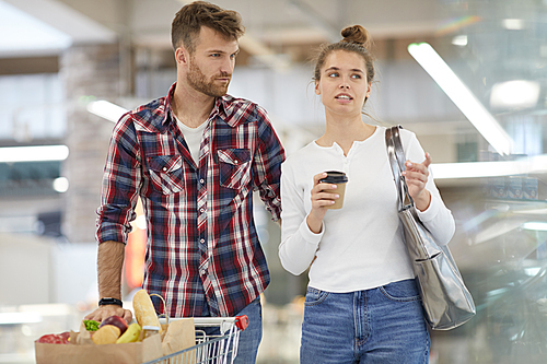 Portrait of contemporary young couple pushing shopping cart while buying groceries in supermarket, copy space