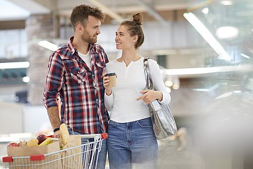 Portrait of contemporary young couple looking at each other and pushing shopping cart while buying groceries in supermarket, copy space