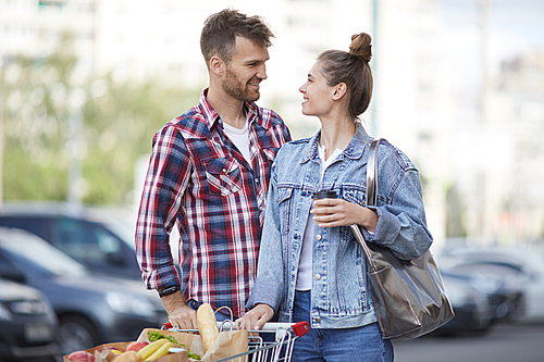 Waist up portrait of contemporary young couple pushing shopping cart with groceries in parking lot, copy space