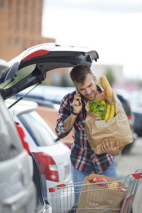 Portrait of handsome young man speaking by phone while packing groceries into car trunk