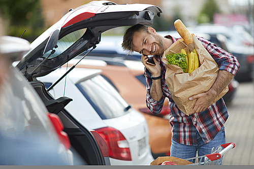 Portrait of smiling young man speaking by phone while packing groceries into car trunk , copy space