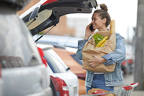 Portrait of smiling young woman speaking by phone while packing groceries into car trunk , copy space