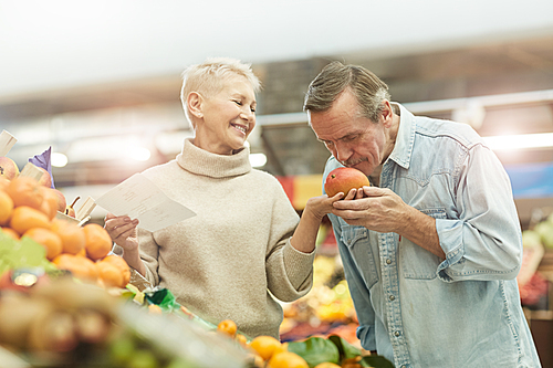 Waist up portrait of modern senior couple smelling fresh fruits while enjoying grocery shopping at farmers market, copy space