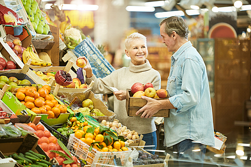 Waist up portrait of smiling senior couple buying fresh fruits while enjoying grocery shopping in farmers market, copy space