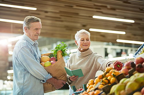 Waist up portrait of smiling senior couple choosing fresh vegetables while enjoying grocery shopping in farmers market, copy space