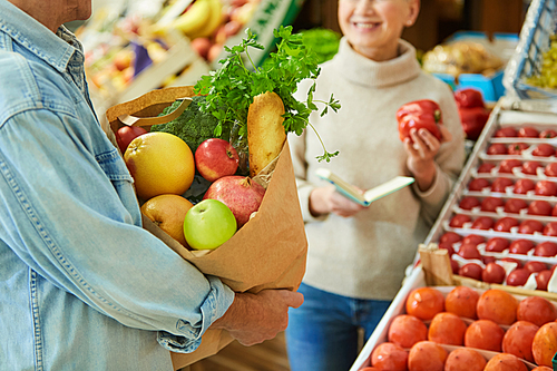 Close up of modern senior couple choosing fresh vegetables while enjoying shopping in farmers market, focus on paper bag with groceries in foreground, copy space