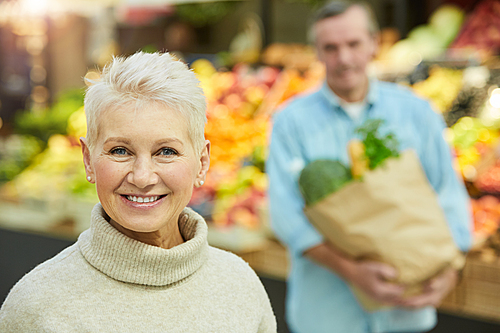 Portrait of modern senior woman holding smiling at camera while enjoying grocery shopping in supermarket with husband in background, copy space