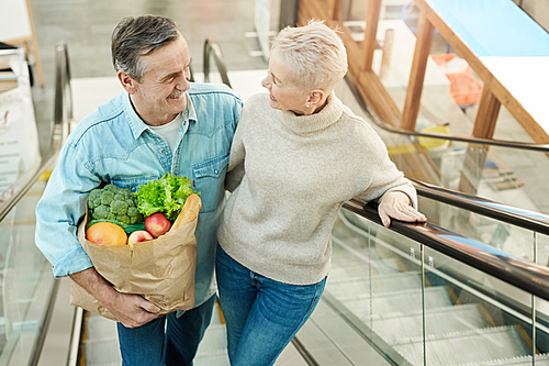 High angle portrait of modern senior couple going up escalator and holding grocery bag while sopping in mall, copy space