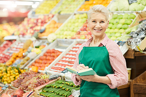 Waist up portrait of smiling senior woman holding notebook while selling fruits and vegetables at farmers market, copy space