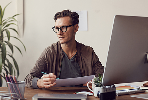 Portrait of handsome businessman wearing glasses holding document while working at desk in modern office, copy space