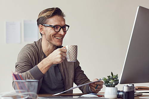 Portrait of handsome adult man smiling cheerfully while looking at computer screen during coffee break in office, copy space