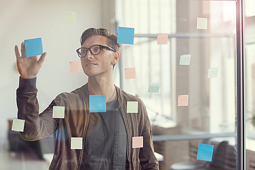 Waist up portrait of handsome adult man planning project and placing colorful stickers on glass wall in office, copy space