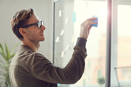 Side view portrait of handsome adult man planning project and placing colorful stickers on glass wall in office, copy space