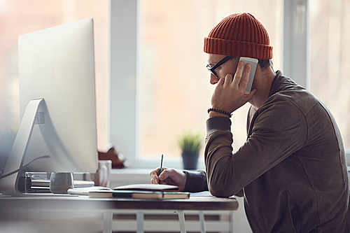 Side view portrait of contemporary businessman wearing glasses and beanie speaking by smartphone while working at desk in office against window, copy space