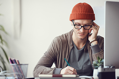 Portrait of contemporary businessman wearing glasses and beanie speaking by smartphone while working at desk in office, copy space
