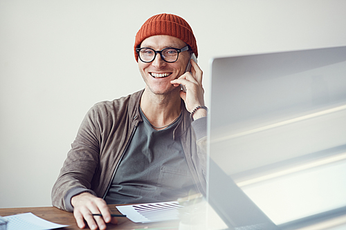 Portrait of contemporary businessman wearing glasses and beanie speaking by smartphone and smiling at camera while working at desk in office, copy space