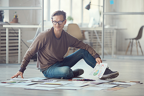 Full length portrait of contemporary businessman sitting on floor in office and planning creative project laying out documents, copy space