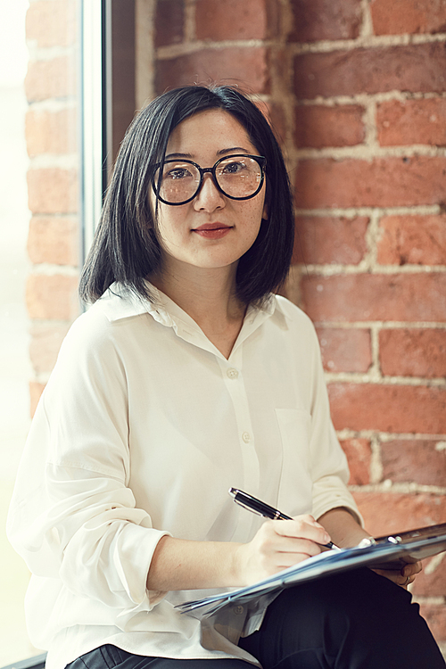 Portrait of successful Asian businesswoman wearing glasses  while writing notes sitting by window in office