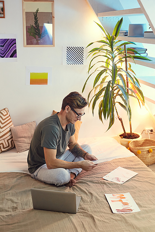 High angle portrait of contemporary man sitting on bed in cozy apartment studio and looking at photographs, copy space