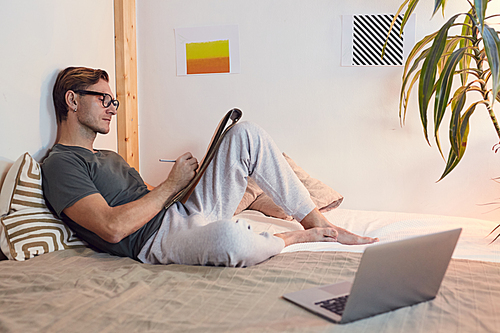 Side view portrait of modern young man lying on bed at home and relaxing by drawing sketches in cozy design interior, copy space
