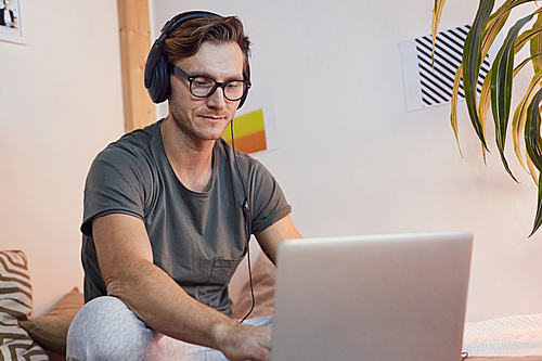 Portrait of contemporary adult man sitting on bed at home and using laptop while wearing big headphones, copy space