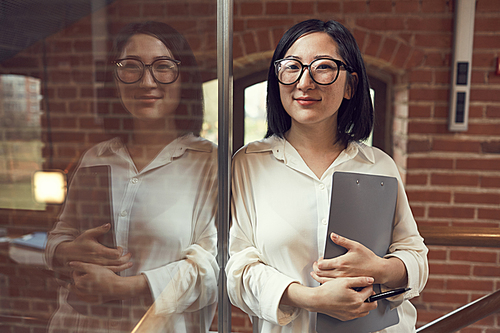 Waist up portrait of young Asian businesswoman wearing glasses smiling at camera while leaning against glass wall with reflection, copy space