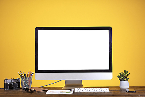 Workplace concept with blank white computer screen at wooden desk over pale yellow background, copy space