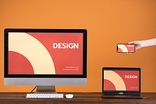 Web design concept with hand holding smartphone, computer and laptop at wooden table over pop orange background, copy space