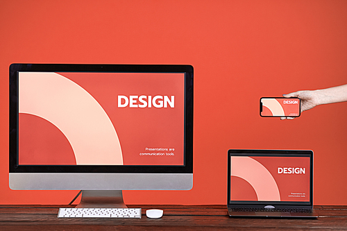 Web design concept with hand holding smartphone, computer and laptop at wooden table over coral background, copy space