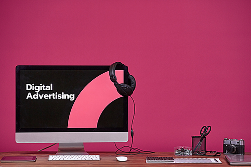 Digital advertising concept with computer and office supplies at wooden table over purple background, copy space