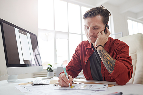 Portrait of contemporary tattooed businessman speaking by phone and taking notes while working at desk in office, copy space