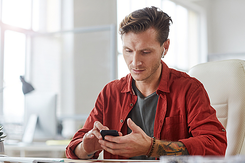 Portrait of contemporary tattooed man using smartphone while working at desk in spacious office, copy space