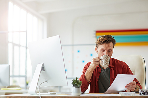 Portrait of modern businessman reading document and drinking coffee while working at desk in spacious white office, copy space