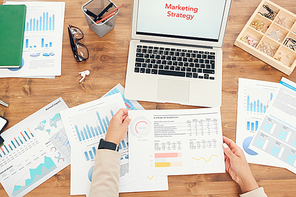 Above view background of unrecognizable businesswoman holding statistics data graph while working at wooden desk with documents laid out and words Marketing Strategy on laptop screen, copy space