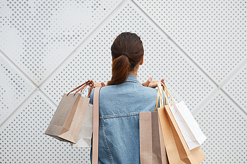 Rear view of young lady with pony tail standing against tiled wall and holding heap of shopping bags