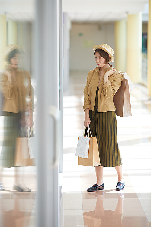 Stylish beautiful lady in straw hat holding many shopping bags and viewing clothes of mannequin through storefront